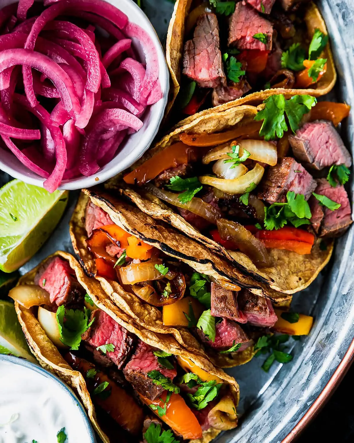 close up view of a platter of steak fajita tacos with pickled onions, limes, and sour cream